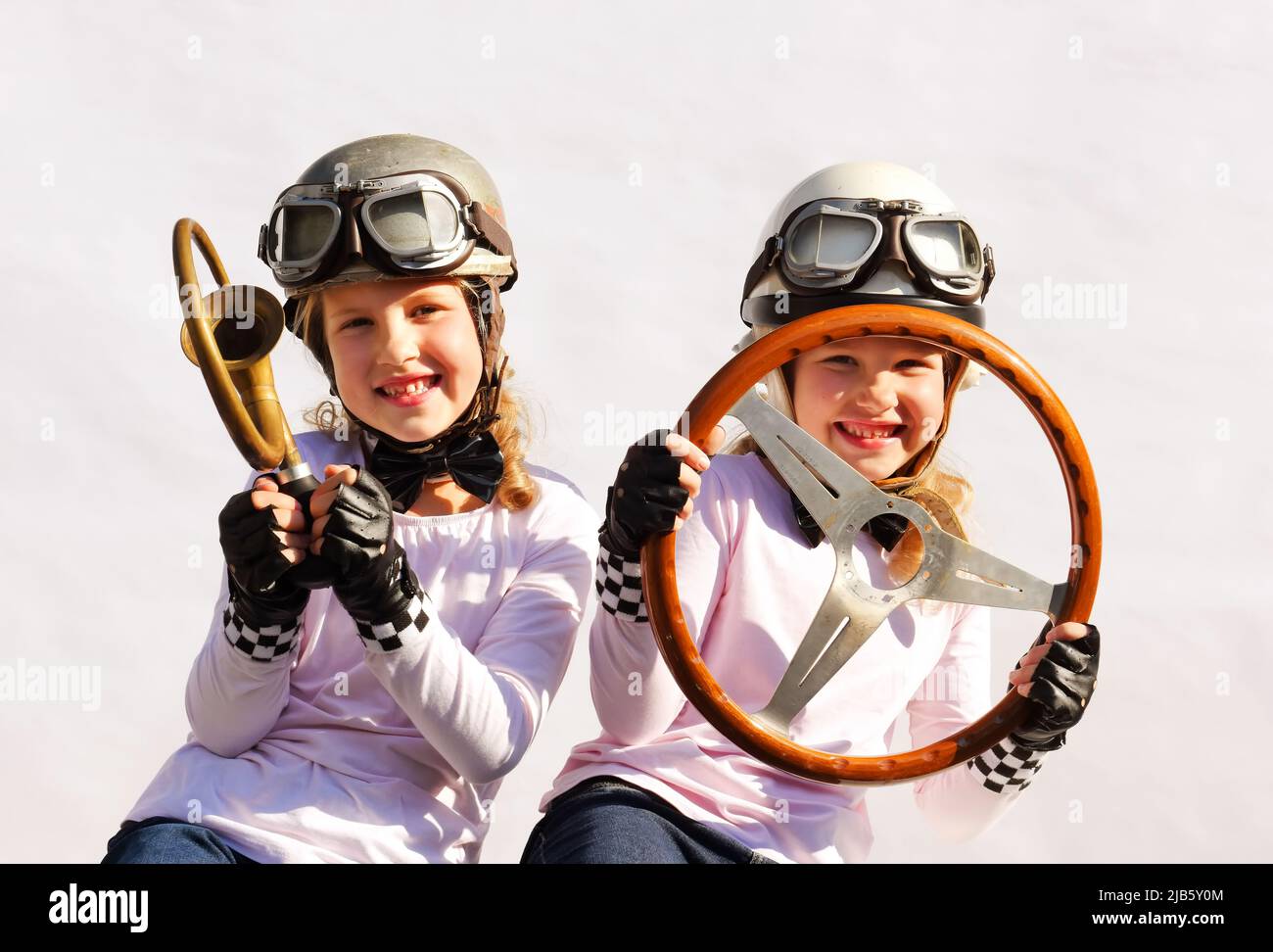 Le sorelle gemelle immaginano che stiano guidando un'auto da corsa. Indossano caschi d'epoca, occhiali e usano un vecchio volante timer e corno per i puntelli. Foto Stock