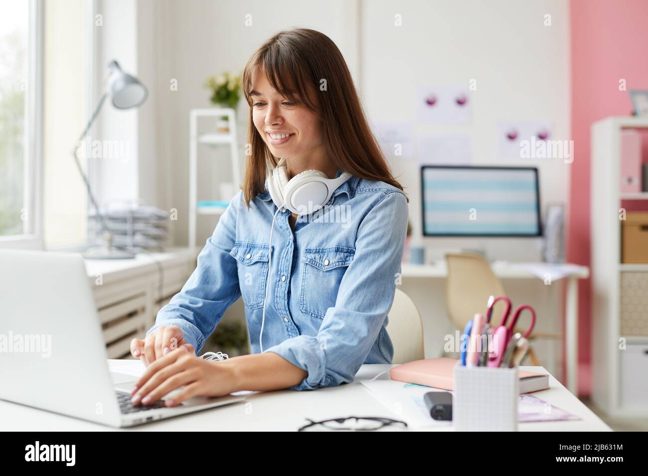 Giovane e ottimista impiegato in un ufficio femminile in denim con cuffie sul collo seduti al tavolo e documenti di editing sul laptop Foto Stock