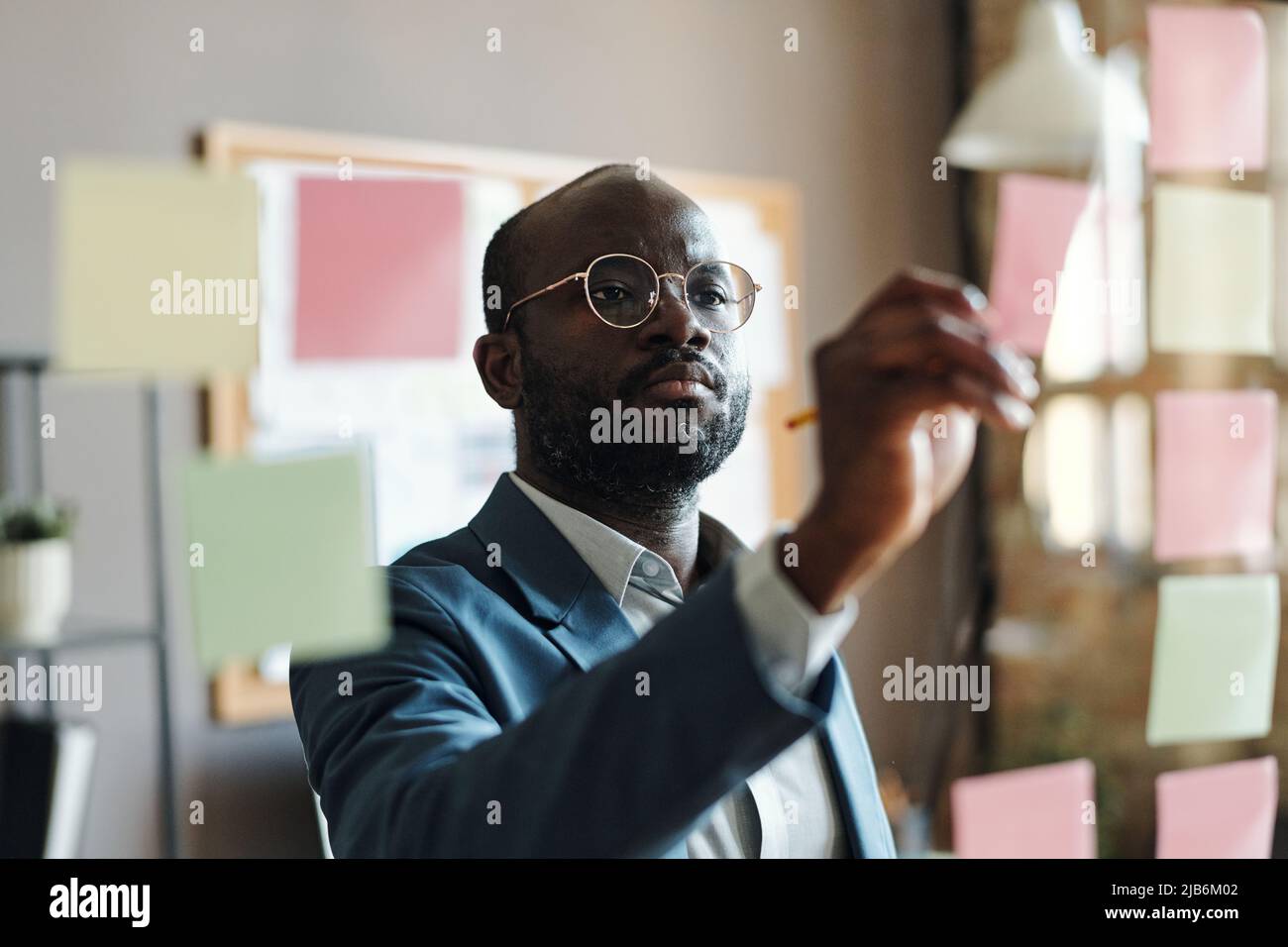 Un uomo d'affari africano serio in vestito e occhiali che fa note su note appiccicose appese su una parete di vetro in ufficio Foto Stock