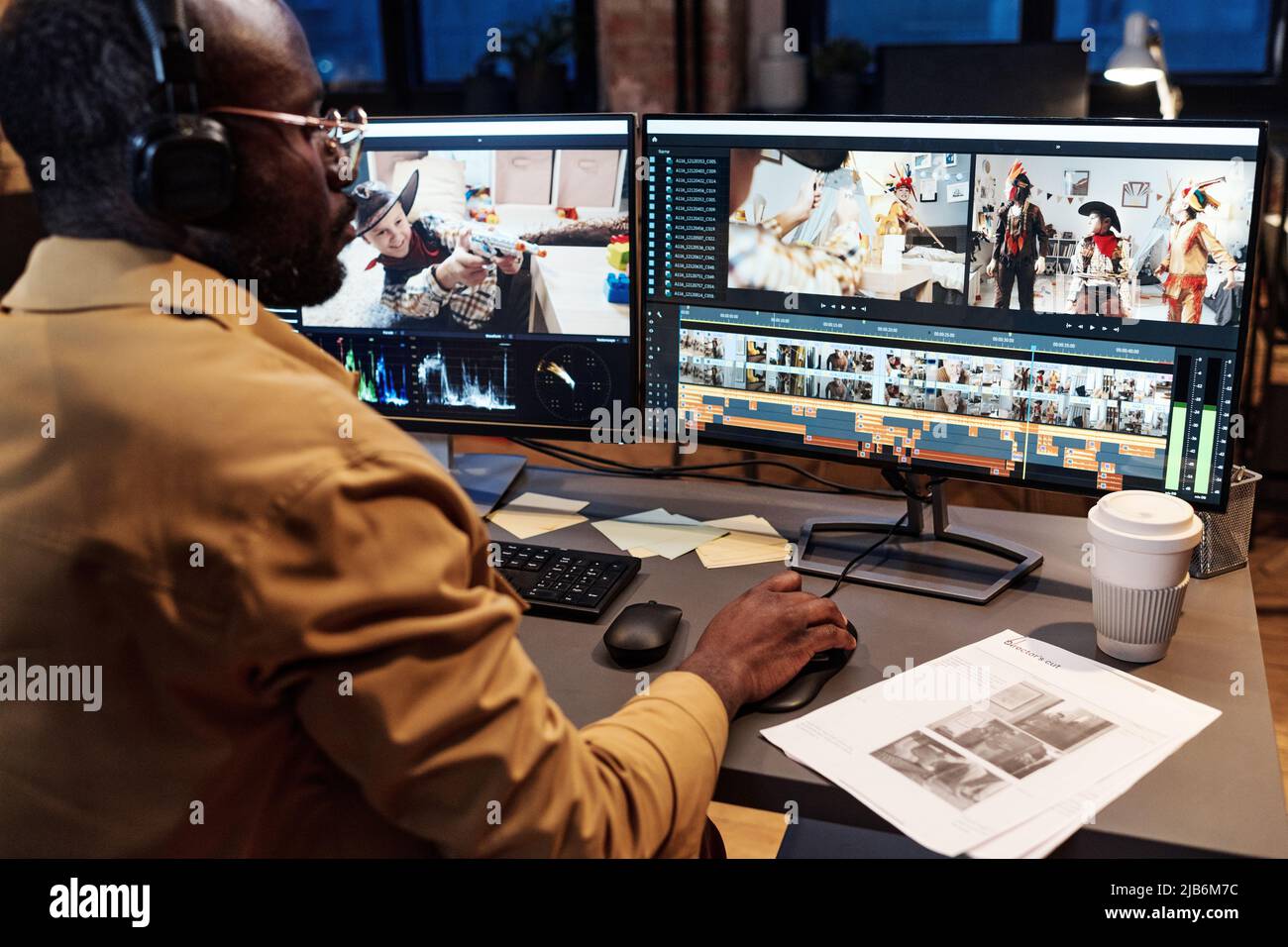 Colorista africano in cuffie guardando il progetto sul tavolo e controllando il lavoro sul monitor del computer al tavolo Foto Stock