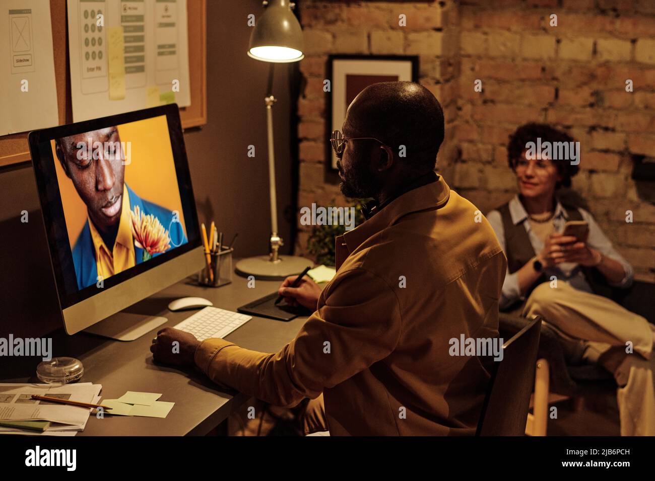 African graphic designer concentrandosi sul suo lavoro al tavolo, guarda il monitor del computer e modifica le foto con un tablet grafico Foto Stock