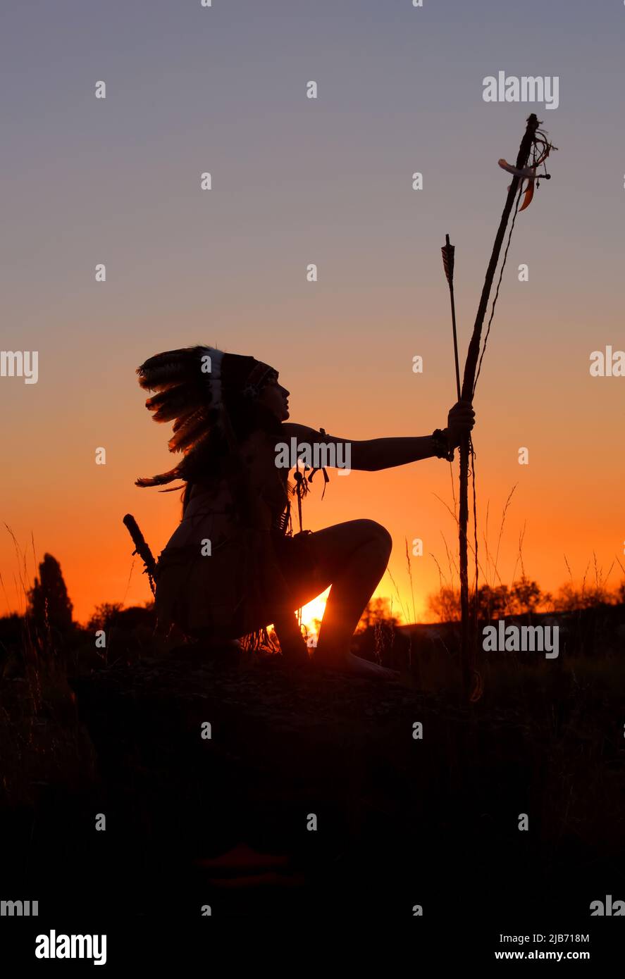 Una ragazza nordamericana Ìndian che indossa un headdress è visto al tramonto. Porta con sé un arco e una freccia. Foto Stock