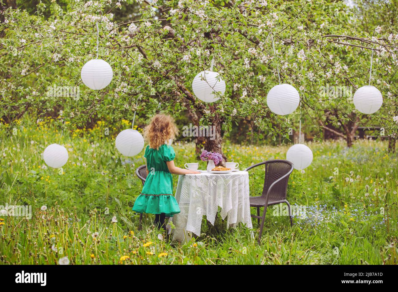 Cute bionda capelli ricci ragazza in piedi in casa Apple giardino da tavolo con té tazze e molto lanterne di carta appeso da fiore albero di mela in primavera. Foto Stock