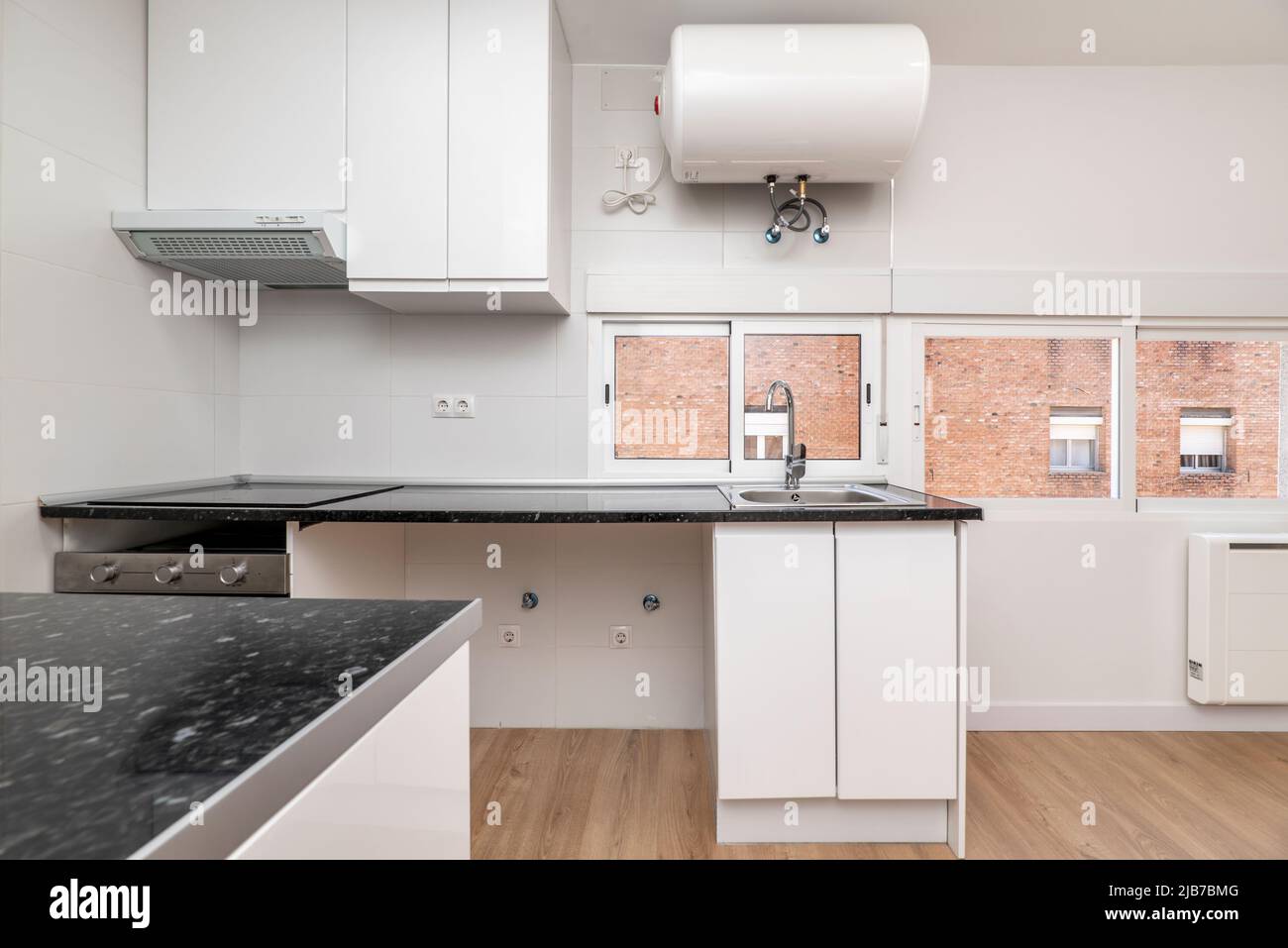 Cucina con armadi bianchi e un piano di lavoro nero lucido con un pavimento in legno duro e finestre lungo una parete, un riscaldatore d'acqua appeso al soffitto Foto Stock