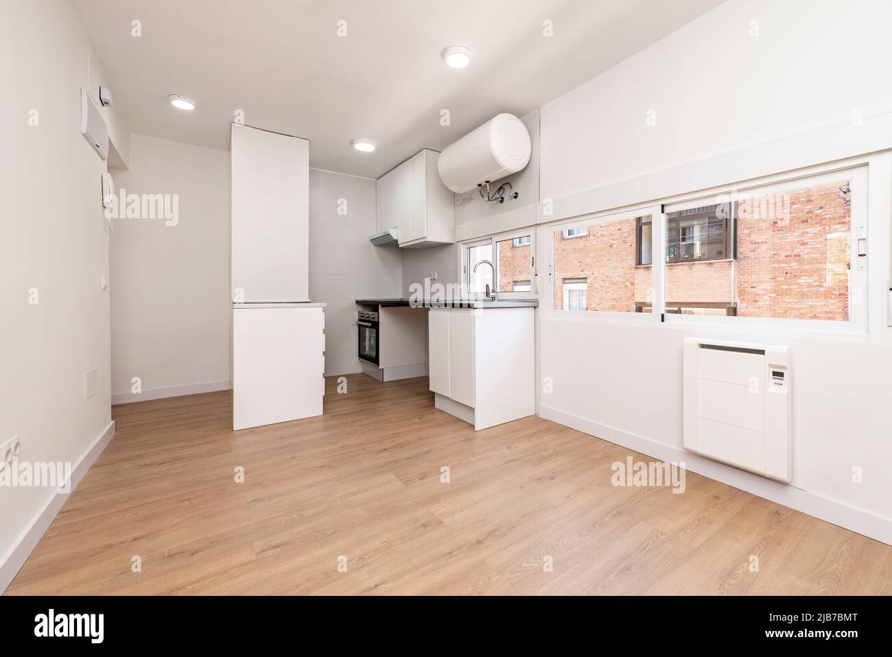 Cucina con armadi bianchi e un piano di lavoro nero lucido con un pavimento in legno duro e finestre lungo una parete, un riscaldatore d'acqua appeso al soffitto Foto Stock