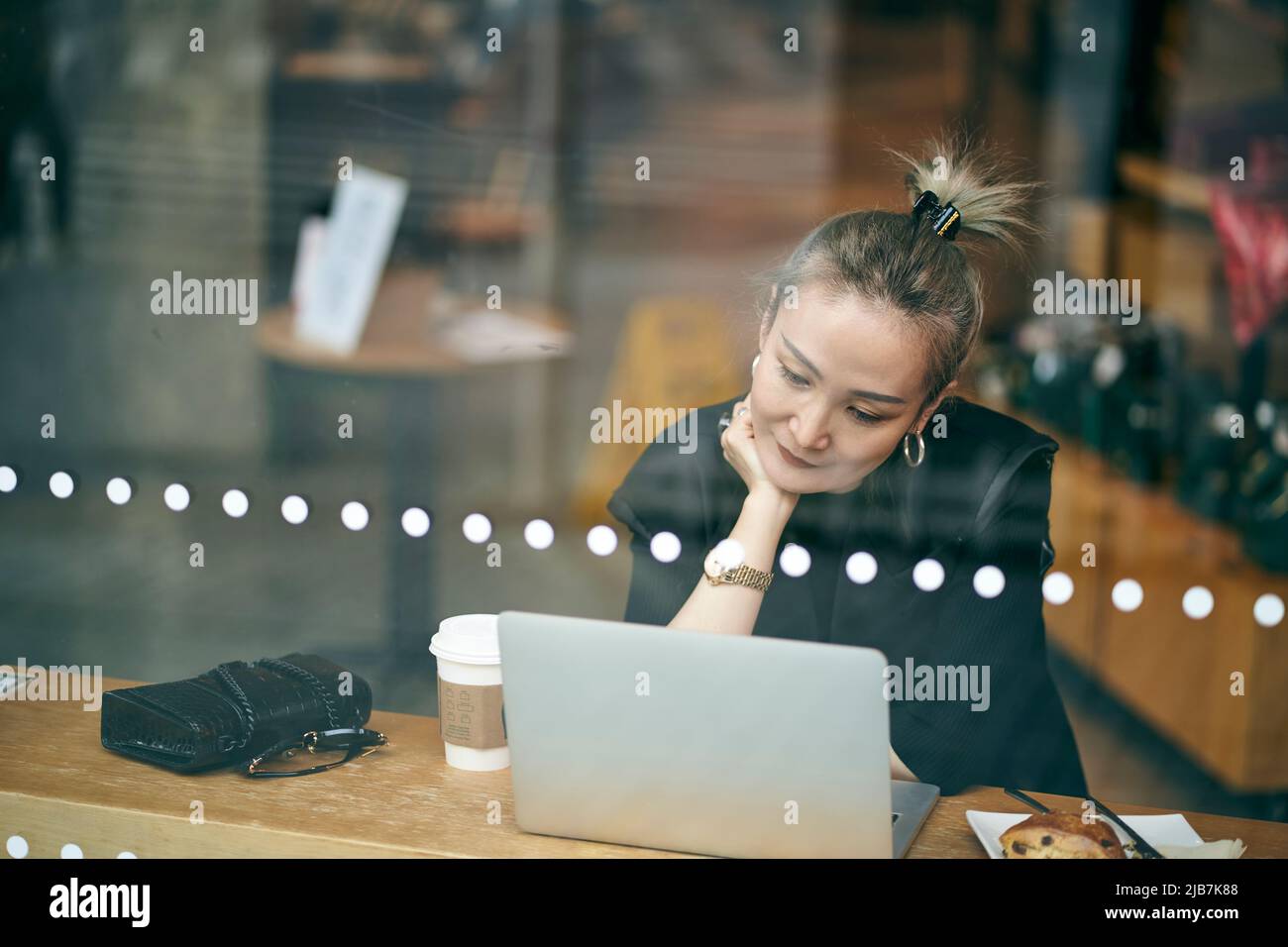 attraverso un colpo di vetro di una donna d'affari asiatica di medio adulto che lavora in un coffee shop utilizzando un computer portatile Foto Stock