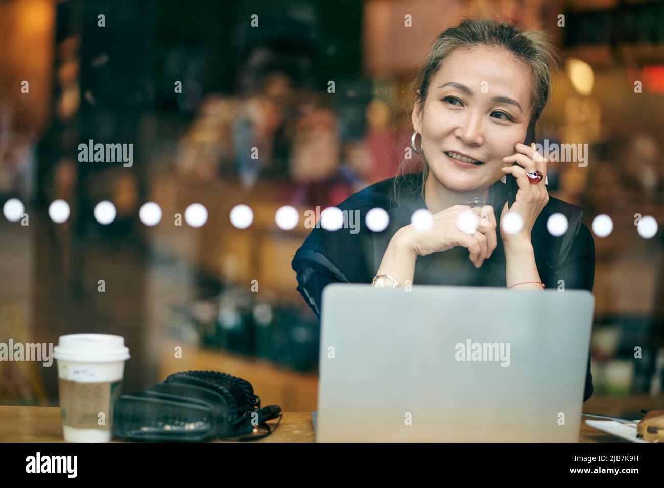 donna d'affari asiatica matura che lavora in caffetteria usando il cellulare e il computer portatile Foto Stock