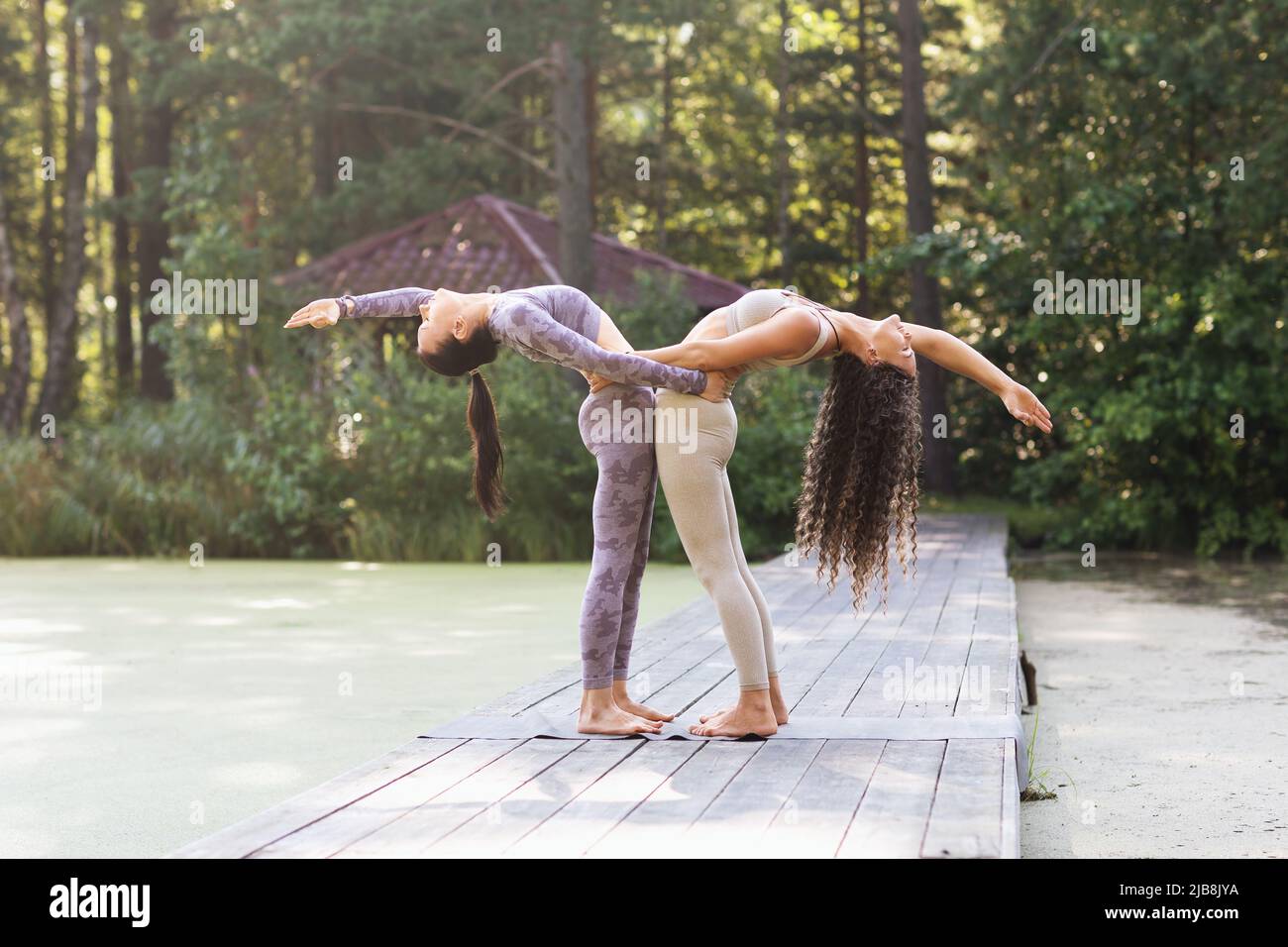 Due praticanti di yoga femminili che si tengono l'un l'altro facendo Hasta Utttanasana, posa in controflessione, in piedi su una passerella di legno in un parco Foto Stock