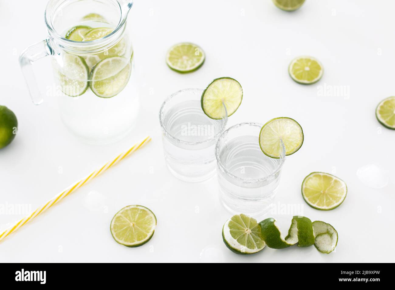 Su un tavolo bianco c'è una brocca di vetro di limonata, accanto ad essa ci sono due bicchieri pieni di limonata e un sacco di lime. Foto Stock