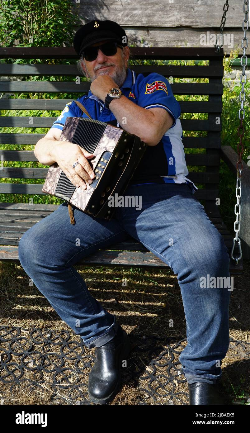L'anziano con il cappello di un capitano si siede all'esterno e tiene una fisarmonica a bottoni diatonica nel braccio. Sembra chiedersi come suonare lo strumento Foto Stock