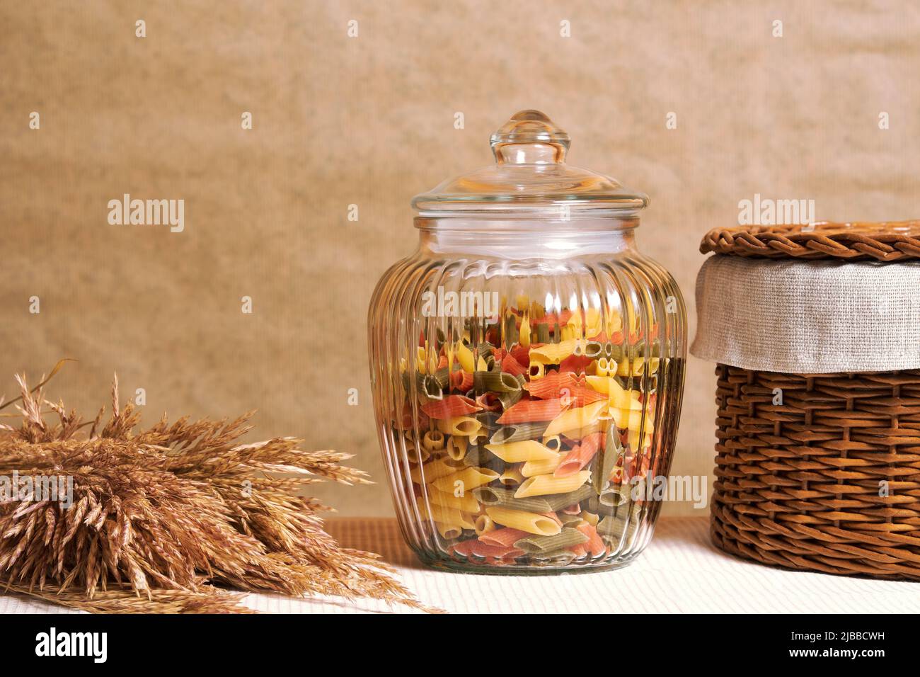 Pasta colorata nel vaso trasparente in cucina con cesto e segale. Sfondo di cibo tradizionale con spazio copia Foto Stock