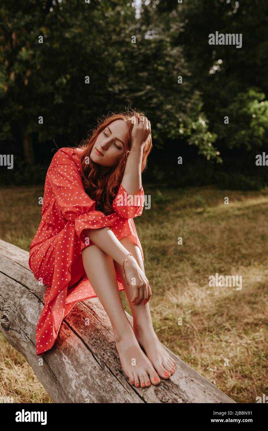 Bella giovane donna adulta in un abito rosso con puntini bianchi con capelli rossi seduti su un albero asciutto caduto e posando su una macchina fotografica. Modella femminile che la tocca Foto Stock