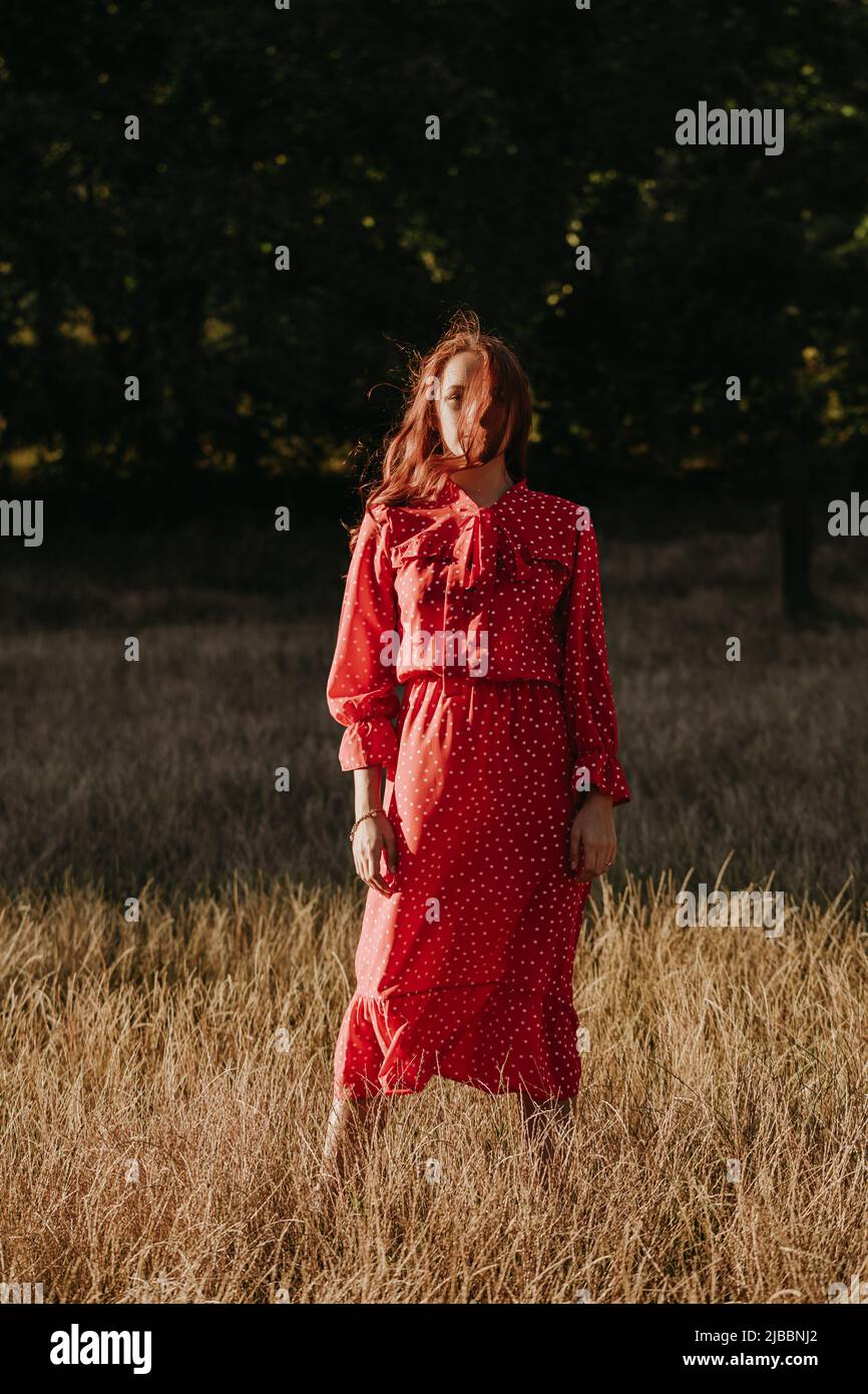 Donna in abito rosso in piedi sul campo sullo sfondo della foresta. Donna che posa su una macchina fotografica alla luce del sole all'aperto Foto Stock