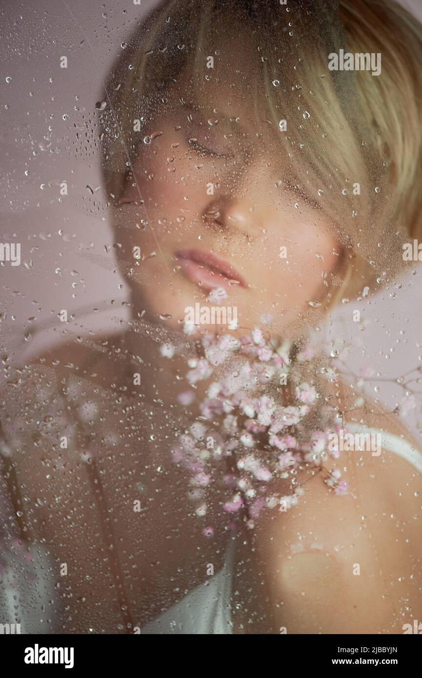 Una ragazza con fiori dietro il vetro bagnato. Nega e divertimento della vita. Occhi chiusi. Industria della bellezza e della cura personale Foto Stock