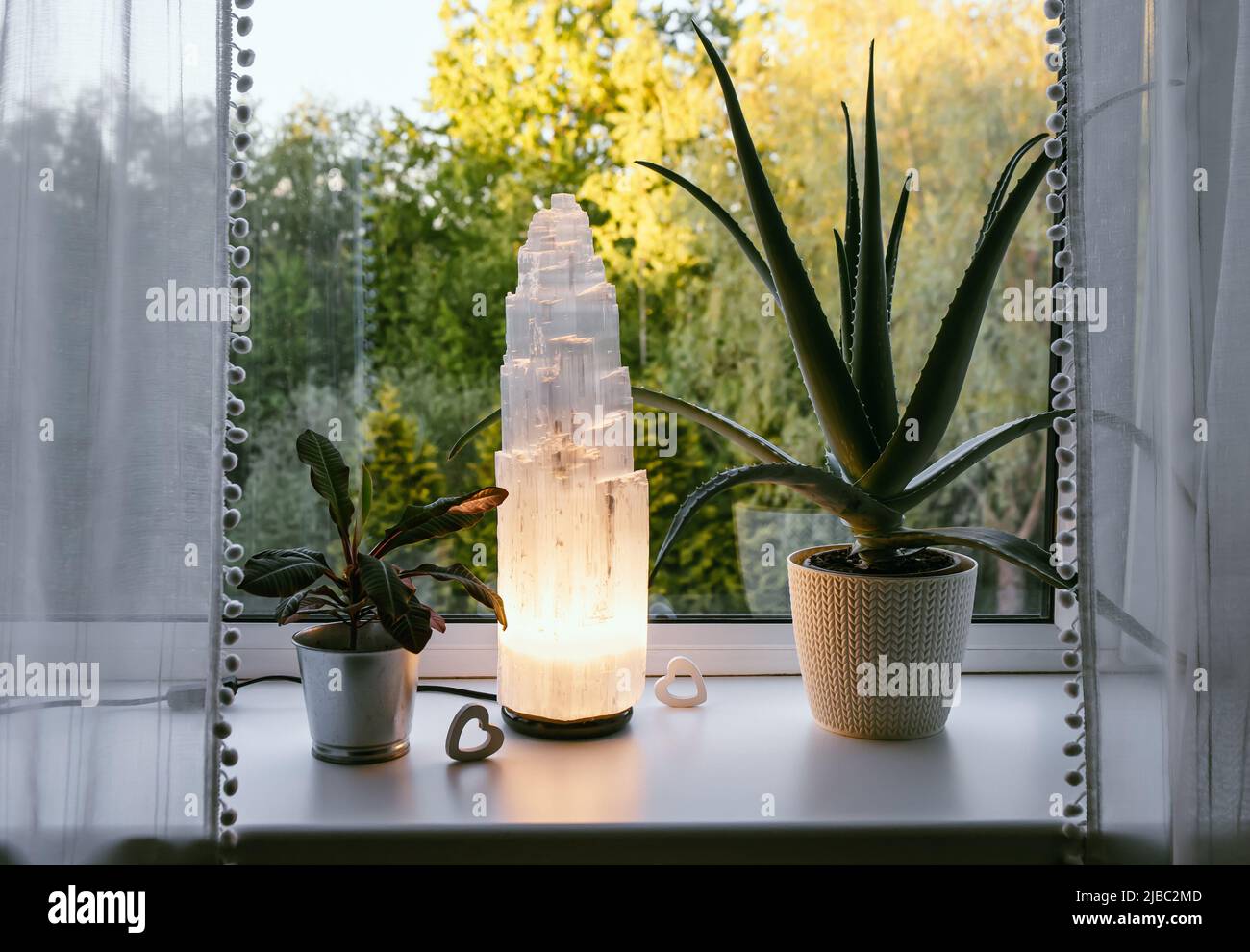 Ruvida grande selenite torre palo lampada illuminata in casa sul davanzale, foresta estiva sullo sfondo, spirituale casa arredamento accento e ambiente mo Foto Stock