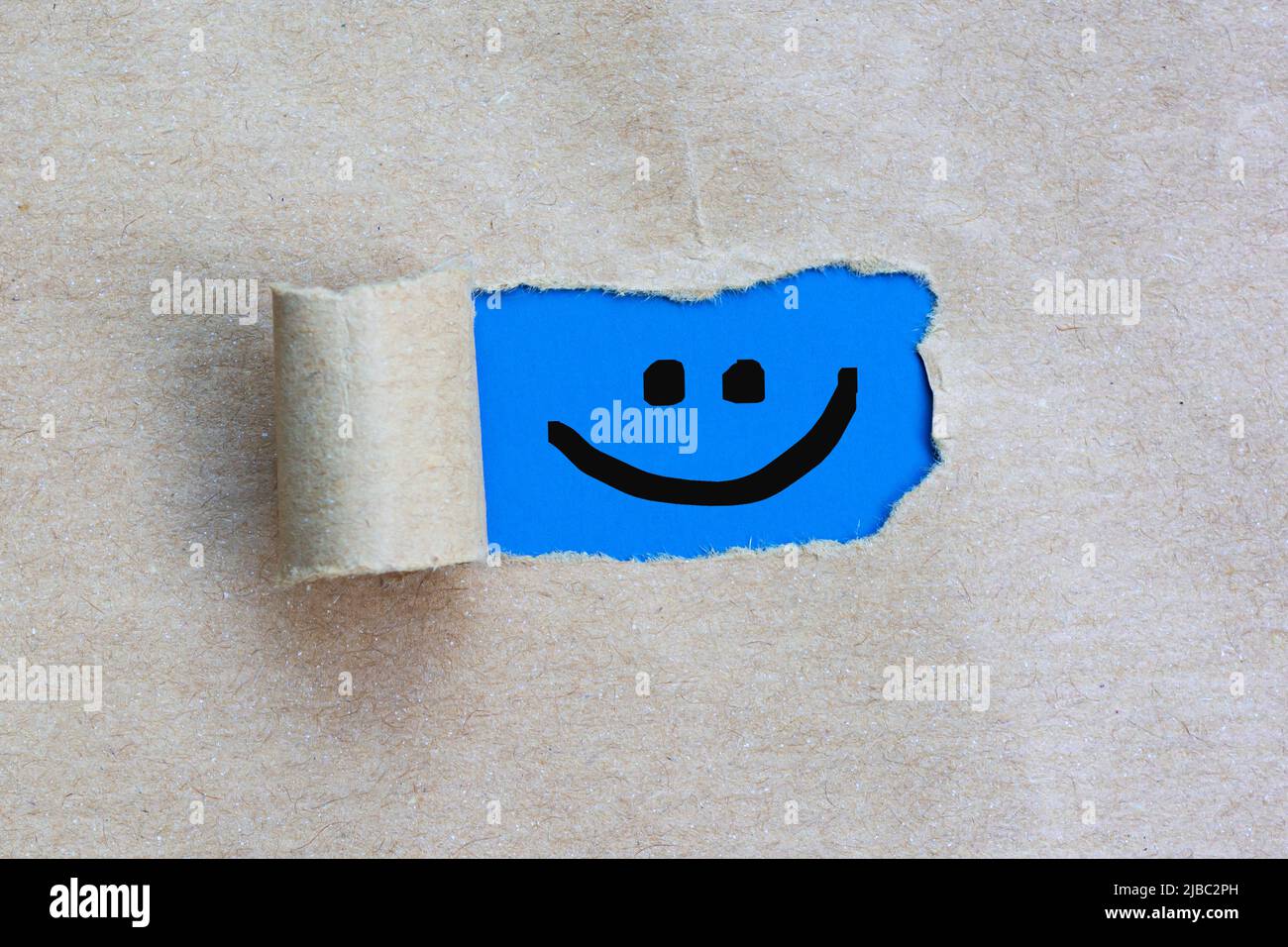 Faccia sorridente felice su buco strappato nel foglio di carta marrone su sfondo blu. Foto Stock