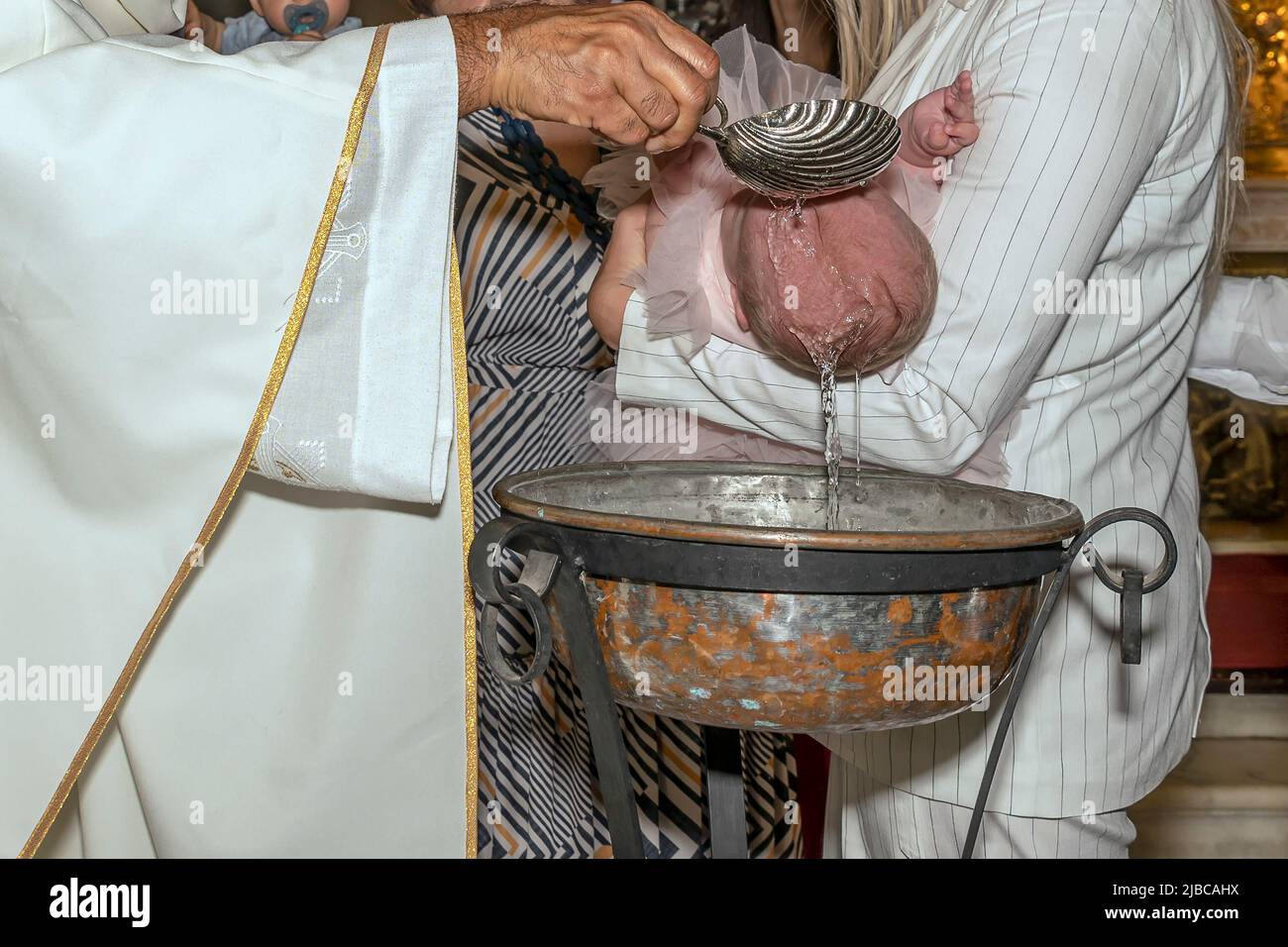 Un neonato viene battezzato nel rito religioso cattolico da un sacerdote vestito di bianco, versando acqua Santa sulla testa Foto Stock