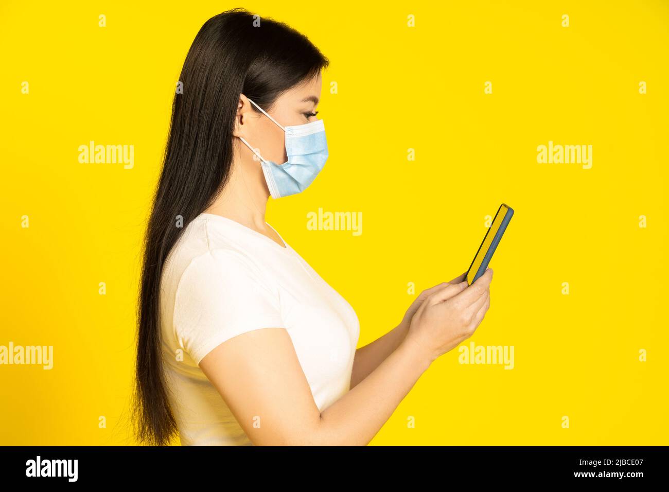 Profilo di mezza lunghezza della donna asiatica di mezza età in maschera medica viso, tenendo smartphone in mano leggere notizie o informazioni importanti. Foto Stock
