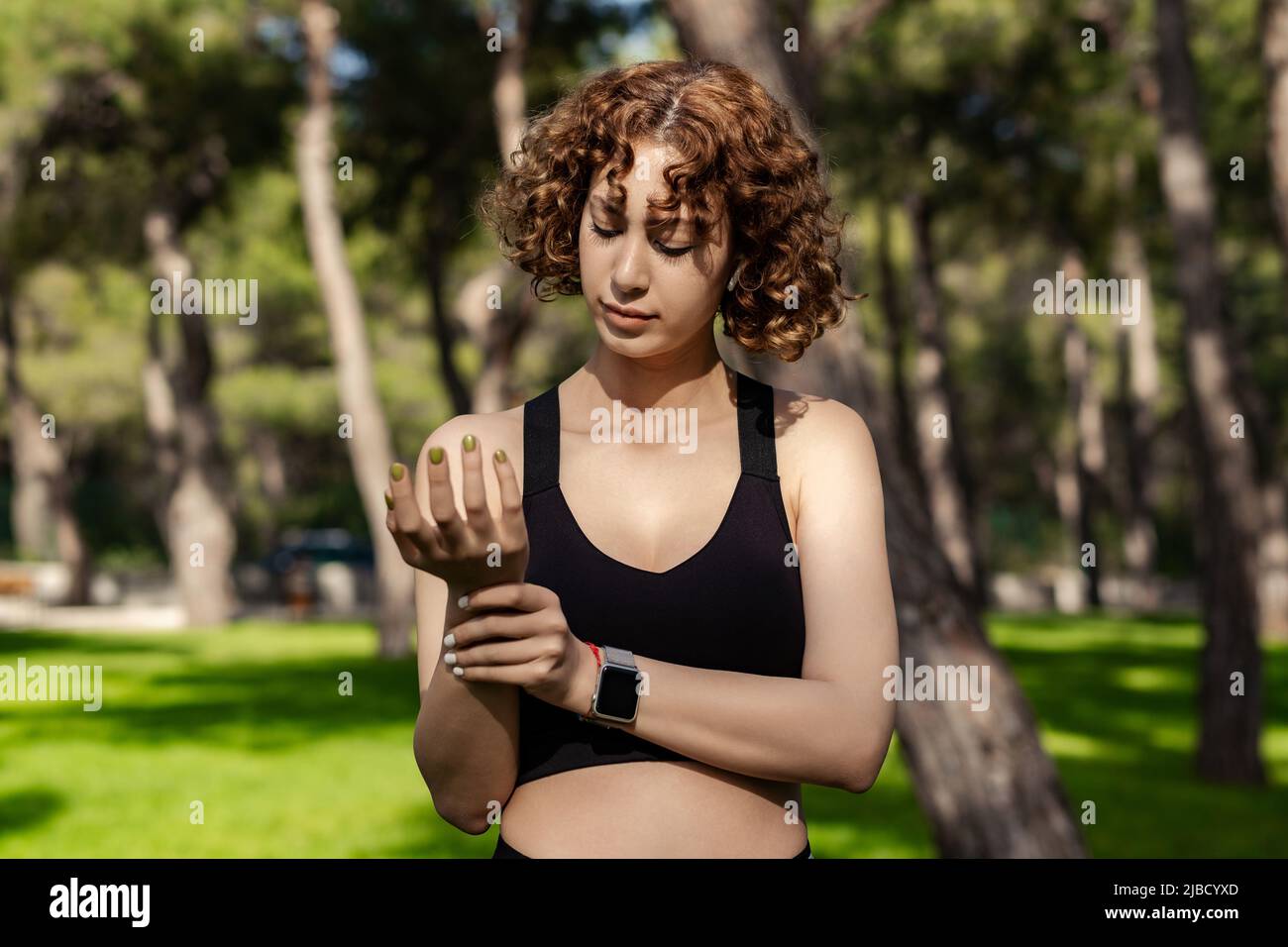 Giovane donna caucasica che indossa un reggiseno sportivo nero in piedi sul parco cittadino, all'aperto tenendo il suo polso doloroso. Dolore alle mani e alle dita, artriti Foto Stock