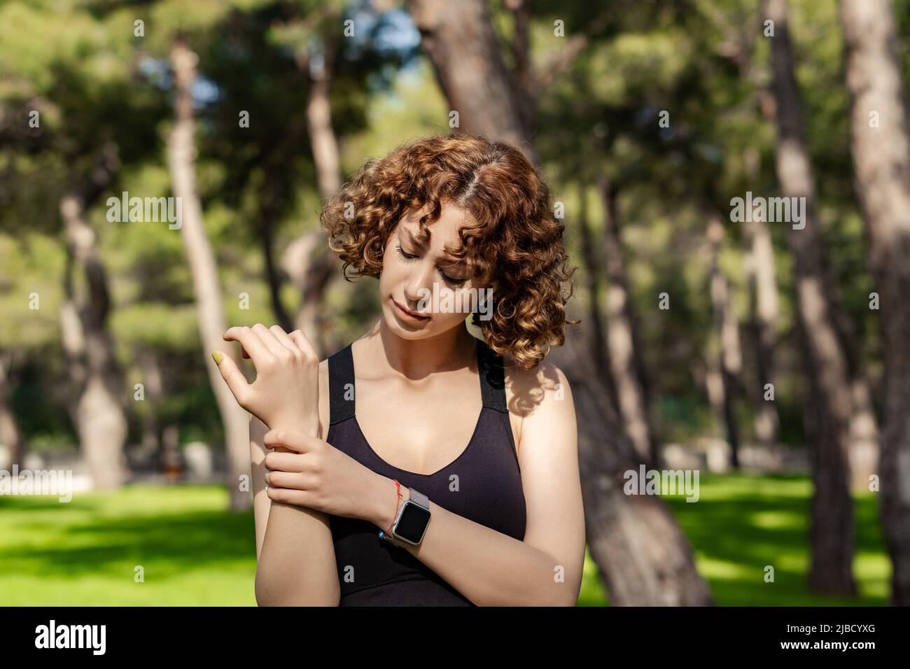 Giovane donna rossa che indossa un reggiseno sportivo nero in piedi sul parco cittadino, all'aperto tenendo il suo polso doloroso. Dolore alle mani e alle dita, artrite Foto Stock