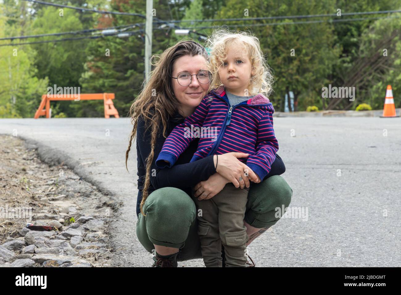 Madre e ragazzo di tre anni stanno in piedi in strada. La mamma ha le braccia avvolte intorno al figlio dopo la tempesta in città. Alberi caduti e coni di traffico sfocati sullo sfondo. Foto Stock