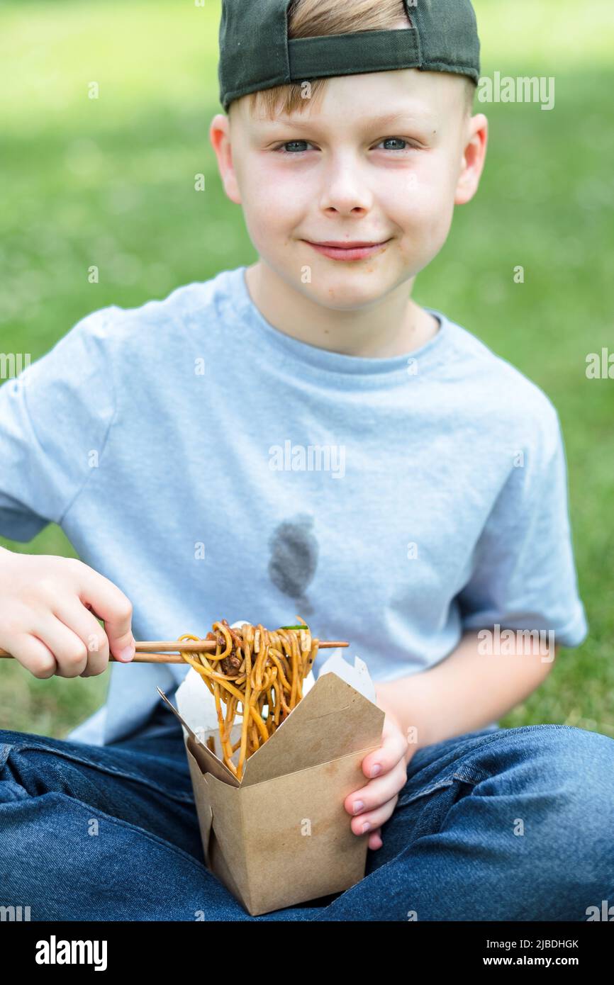 Il ragazzo in mano reggendo un bastone con salsa di soia spaghetti fritti in padella. Sporco macchia di salsa sui suoi vestiti. All'aperto Foto Stock