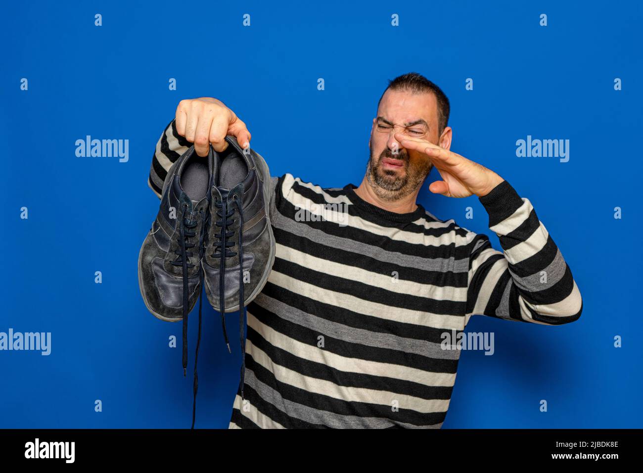 Un uomo tiene le sneakers nelle sue mani. Odore sgradevole. Gambe stinky, sudorazione eccessiva dei piedi. Isolato su sfondo blu studio. Foto Stock