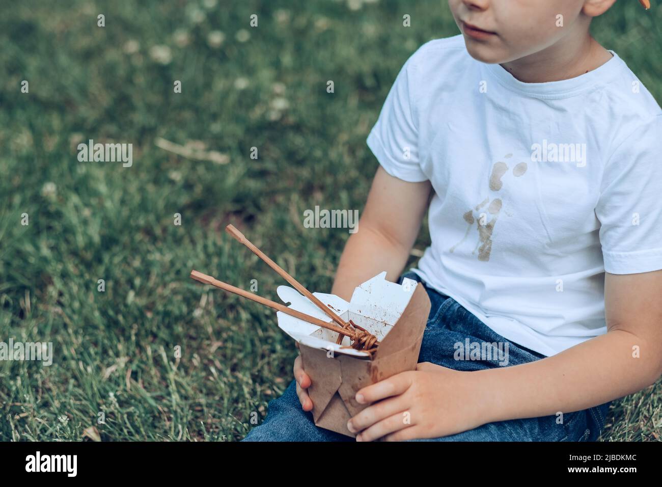 Il ragazzo che tiene il cibo da asporto seduto sull'erba. La salsa sporca si macchia su una maglietta bianca. All'aperto Foto Stock