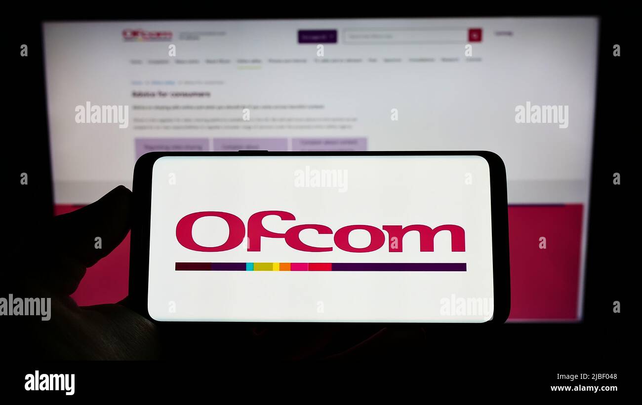 Persona che tiene sullo schermo il cellulare con il logo dell'ufficio delle comunicazioni dell'autorità britannica (UFCOM) davanti alla pagina web. Mettere a fuoco sul display del telefono. Foto Stock