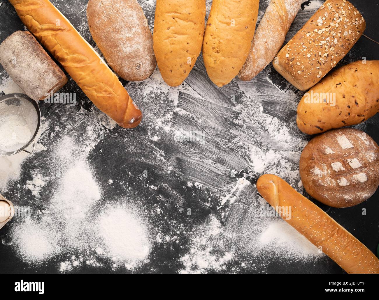 un vasto assortimento di diversi tipi di pane disposti su sfondo nero con farina, dolci caldi freschi, farina di grano, produzione di pane Foto Stock
