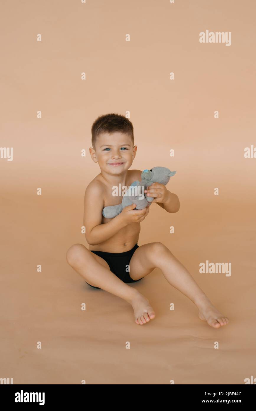 Un bambino in mutande tiene in mano un morbido giocattolo dell'orso, seduto su sfondo beige in studio e sorridente Foto Stock