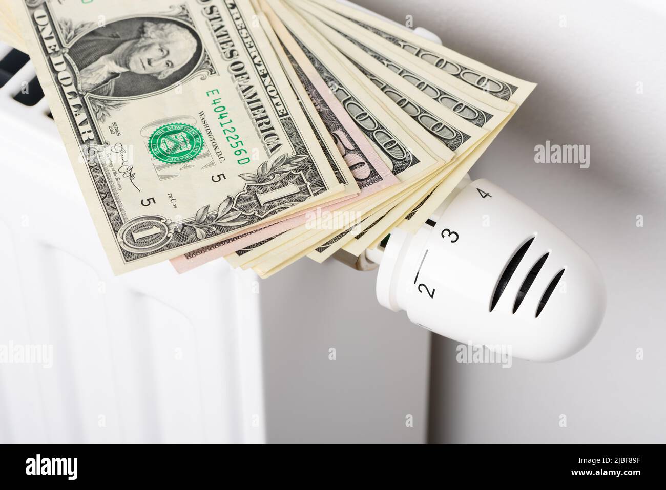 Pila di banconote in dollari sulla batteria del radiatore di riscaldamento con termostato regolatore di temperatura. Concetto di costi di riscaldamento costosi e aumento della bolletta energetica Foto Stock