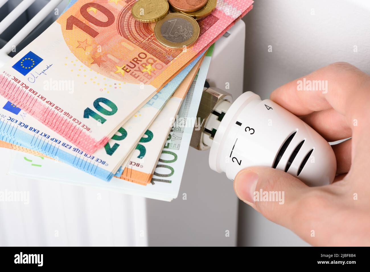 Regolazione manuale della manopola della valvola del termostato di temperatura del radiatore di riscaldamento con pila di banconote in euro. Costi di riscaldamento costosi e in aumento Foto Stock