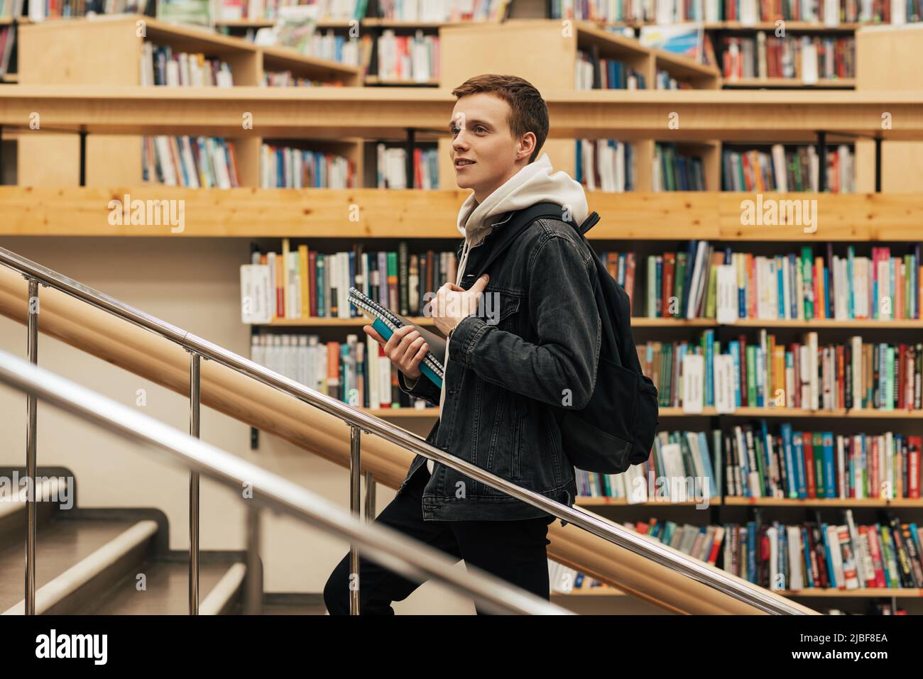 Giovane studente maschio salendo le scale in biblioteca. Uomo con libri di testo in biblioteca universitaria. Foto Stock