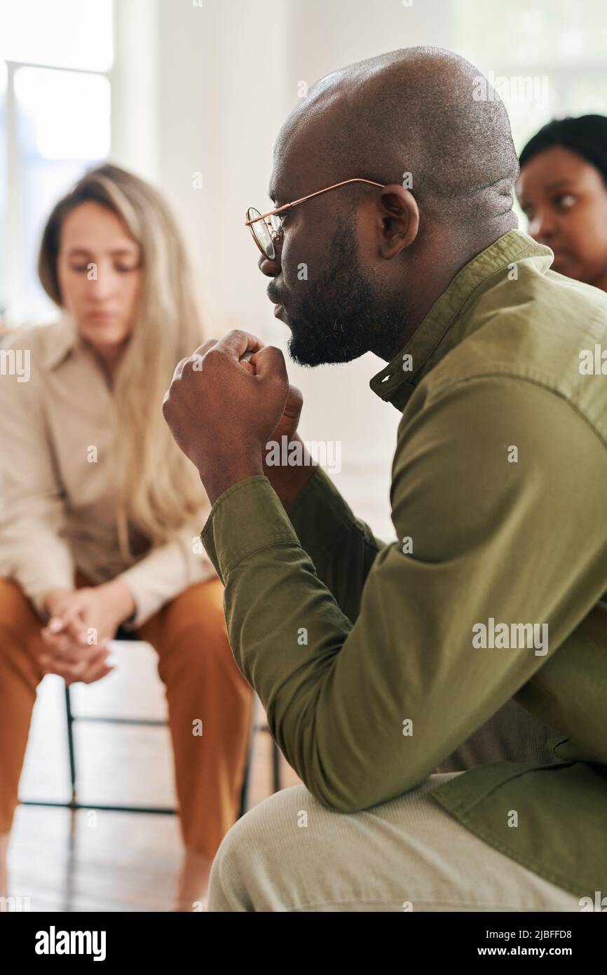 Vista laterale del giovane uomo stressato con le mani interbloccate dal suo volto che condivide il suo problema durante la sessione psicologica Foto Stock