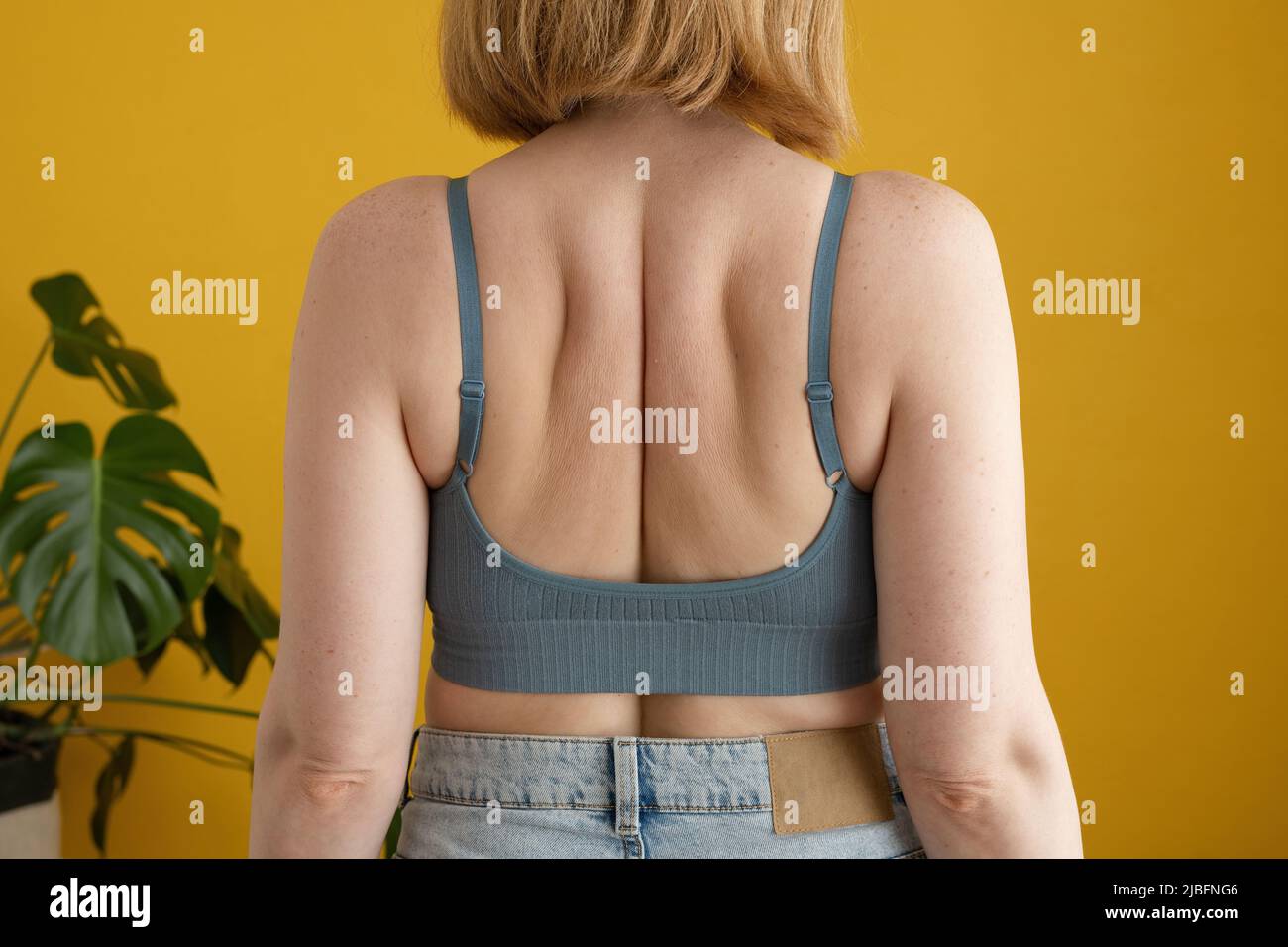 Vista posteriore di una donna irriconoscibile con capelli zenzero e corpo curvo in piedi contro il muro giallo in casa Foto Stock
