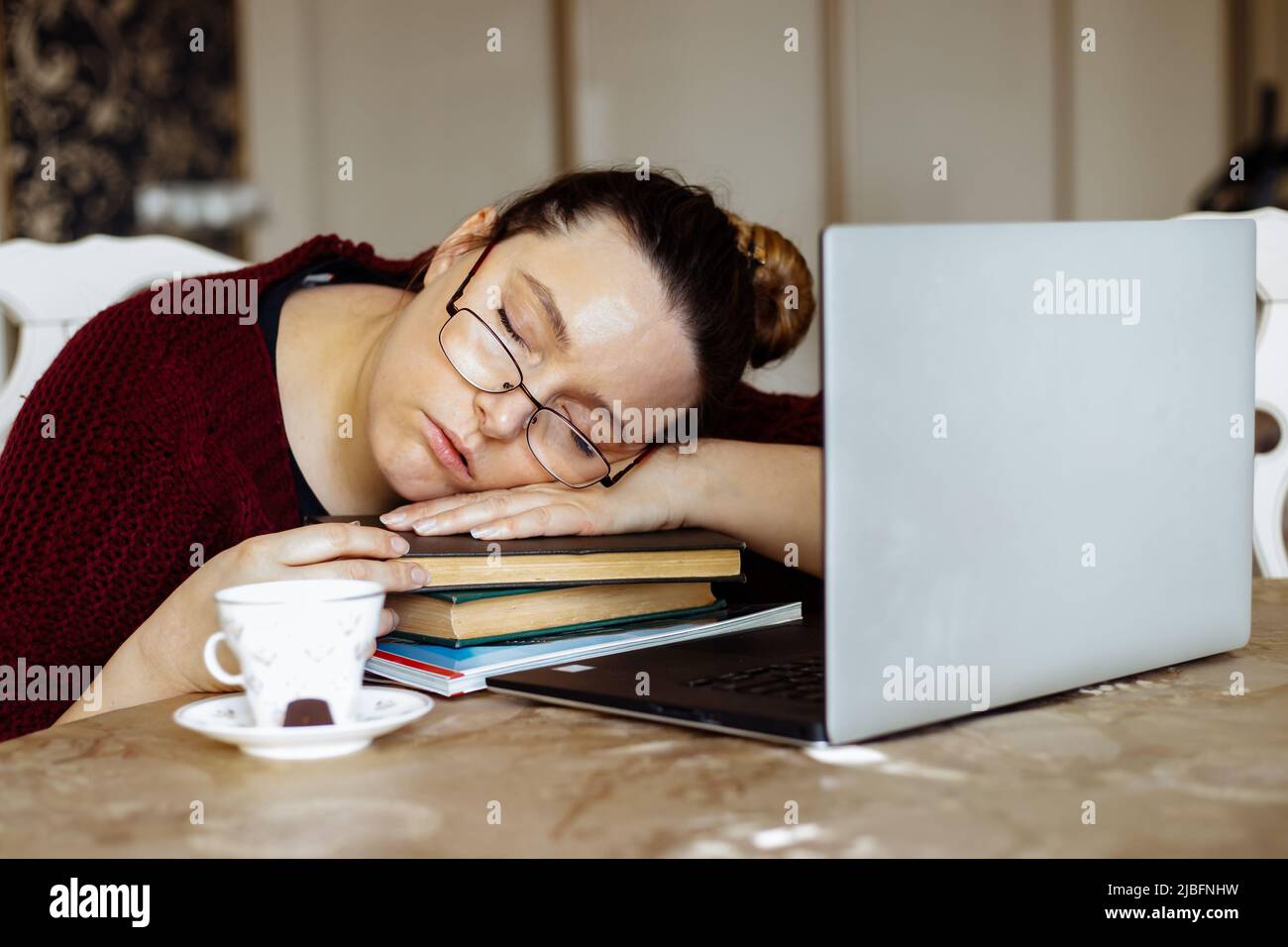 Ritratto di esausta donna di mezza età seduta a tavola vicino a tazza, laptop, dormire su braccia incrociate su mucchio di libri. Foto Stock