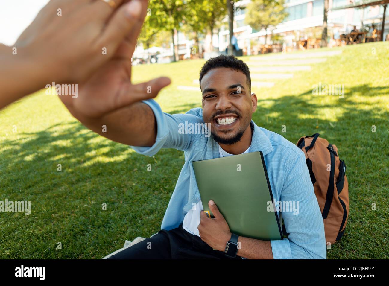 Felice afroamericano che dà cinque alti, celebrando il successo accademico e superato l'esame, seduto sul prato all'aperto Foto Stock
