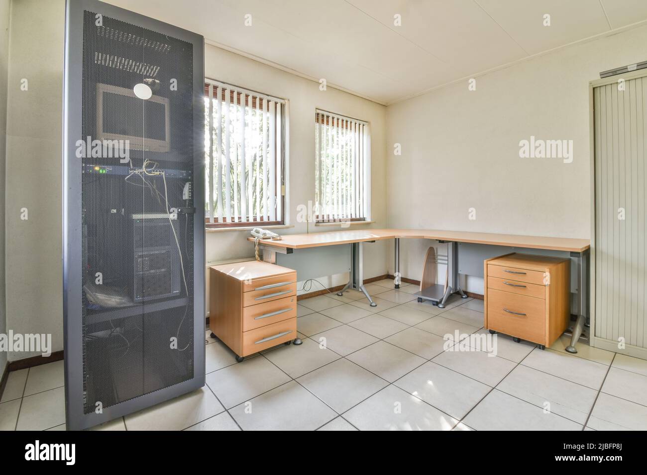 Computer in cabinet con porta a rete vicino a tavolo di legno con telefono posto a finestre con jalousie in ufficio con pareti bianche e pavimento piastrellato Foto Stock