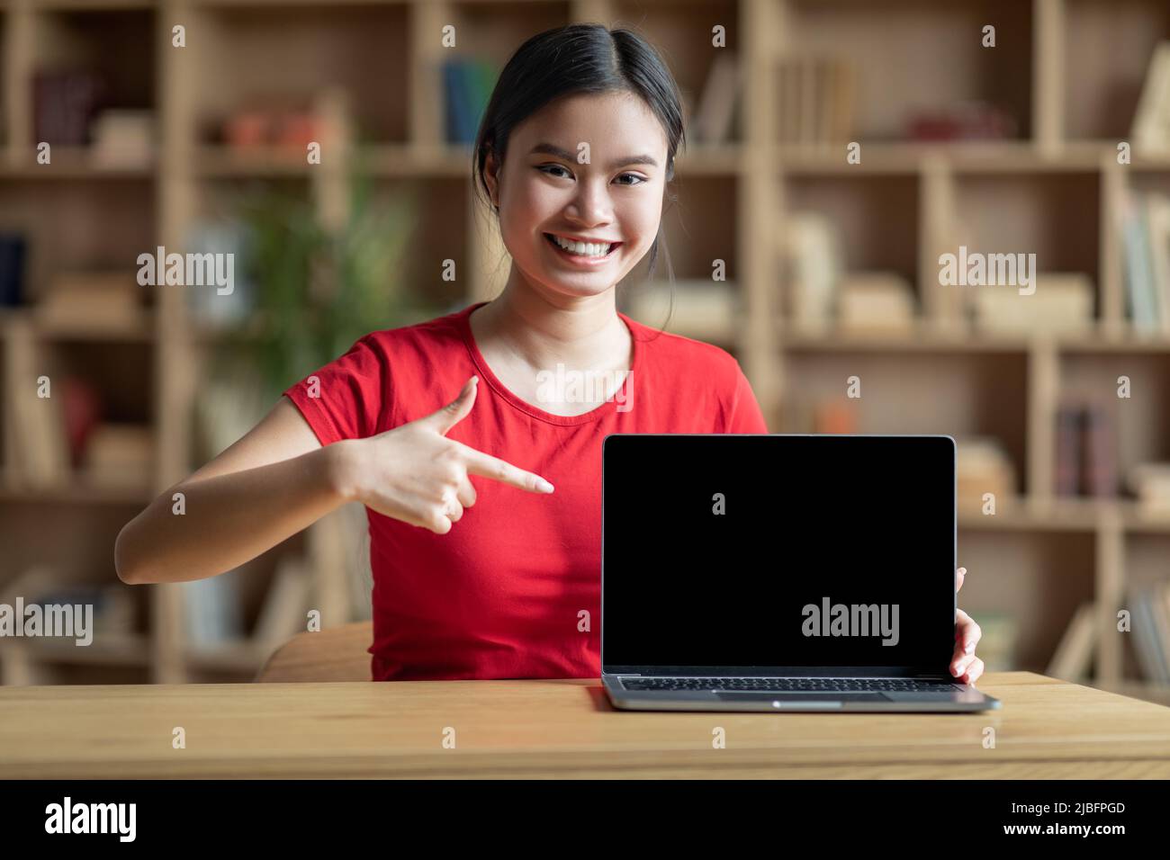 Sorridente donna coreana punta il dito al computer con schermo vuoto per lo studio in camera interna Foto Stock
