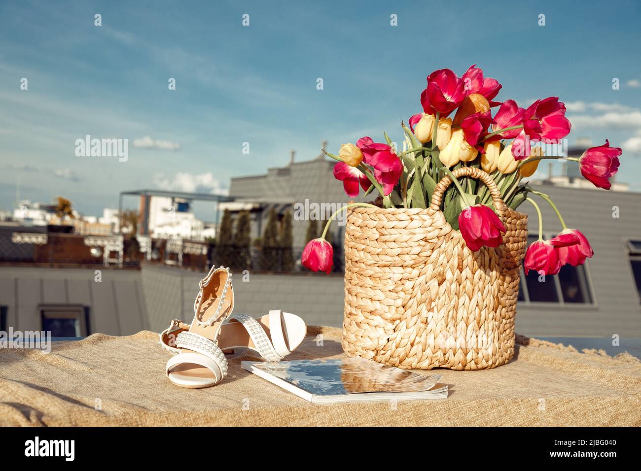 Composizione di sacchetto di paglia con mazzo di tulipani rosa, giallo fiori, sandali beige e rivista su tela di lino sul tetto. Foto Stock