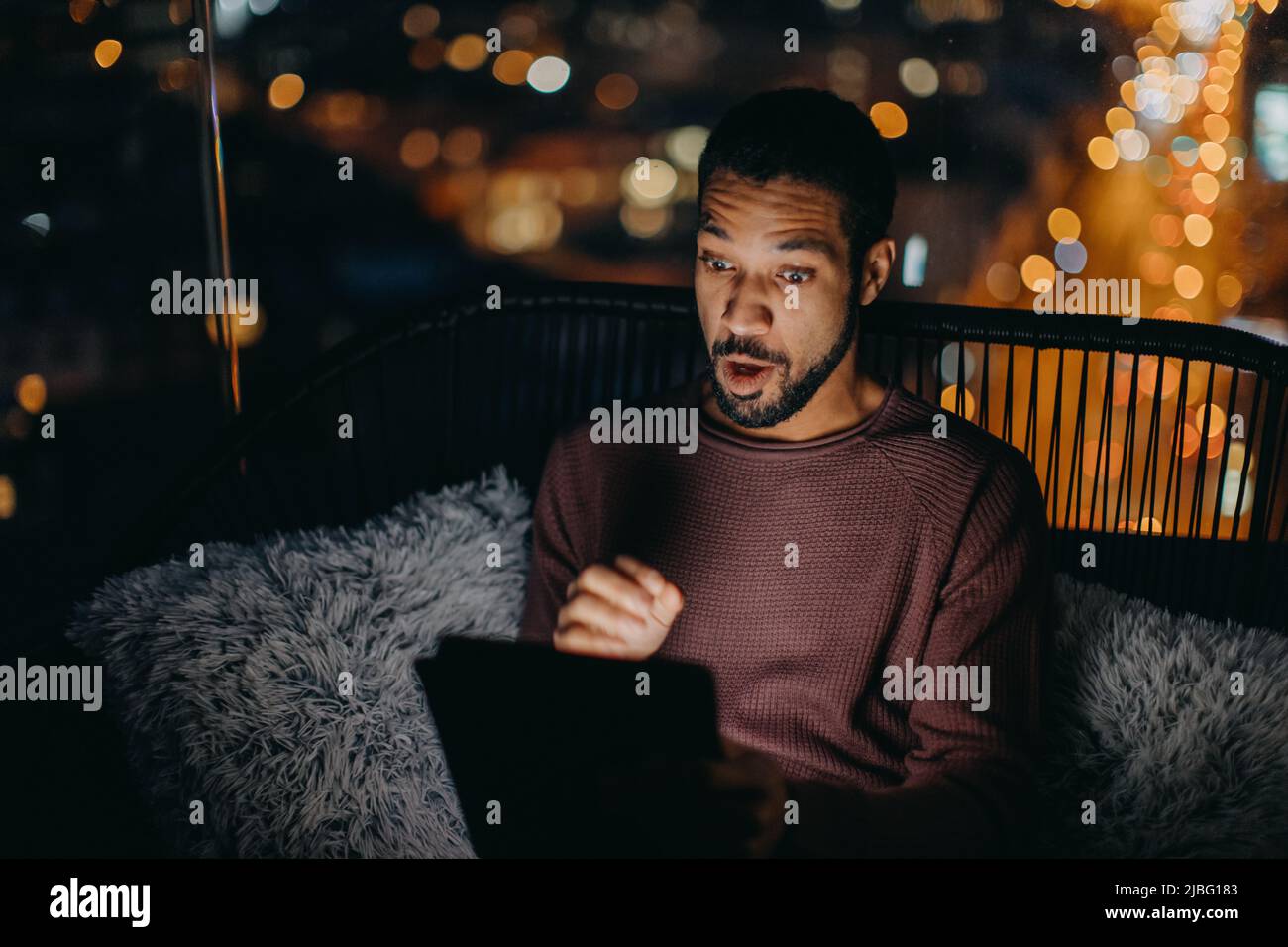 Giovane afroamericano seduto sul balcone con vista sulla città e con un tablet di notte Foto Stock