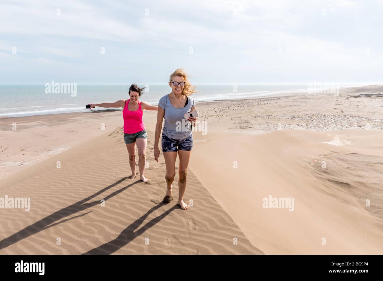 Vista frontale di due donne che camminano sulla cima di una grande duna, una di loro con le braccia aperte al vento. Madre e figlia Foto Stock