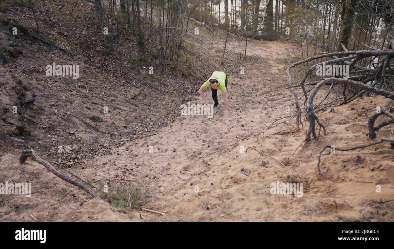 L'uomo giovane durante la corsa salta su ripide pendii senza fermarsi. Runner in t-shirt giallo brillante e cuffie si arrampica sulla collina di sabbia con l'aiuto delle mani Foto Stock