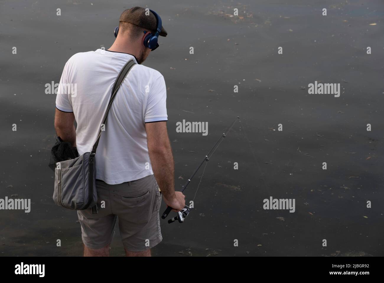Il giovane uomo in camicia bianca, berretto, cuffie e borsa a spalla è pesca con una canna da pesca Foto Stock