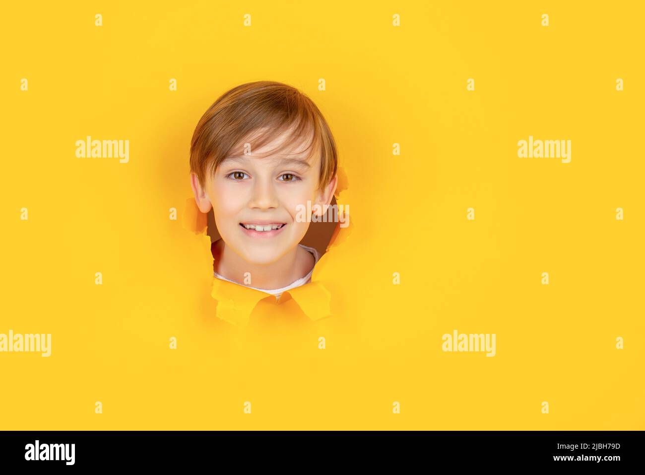 Il capretto con il sorriso toothy mostra la faccia nel foro di carta. Bambino positivo con un sorriso piacevole sul viso, si tiene attraverso il buco lacerato in carta blu. Rottura Foto Stock