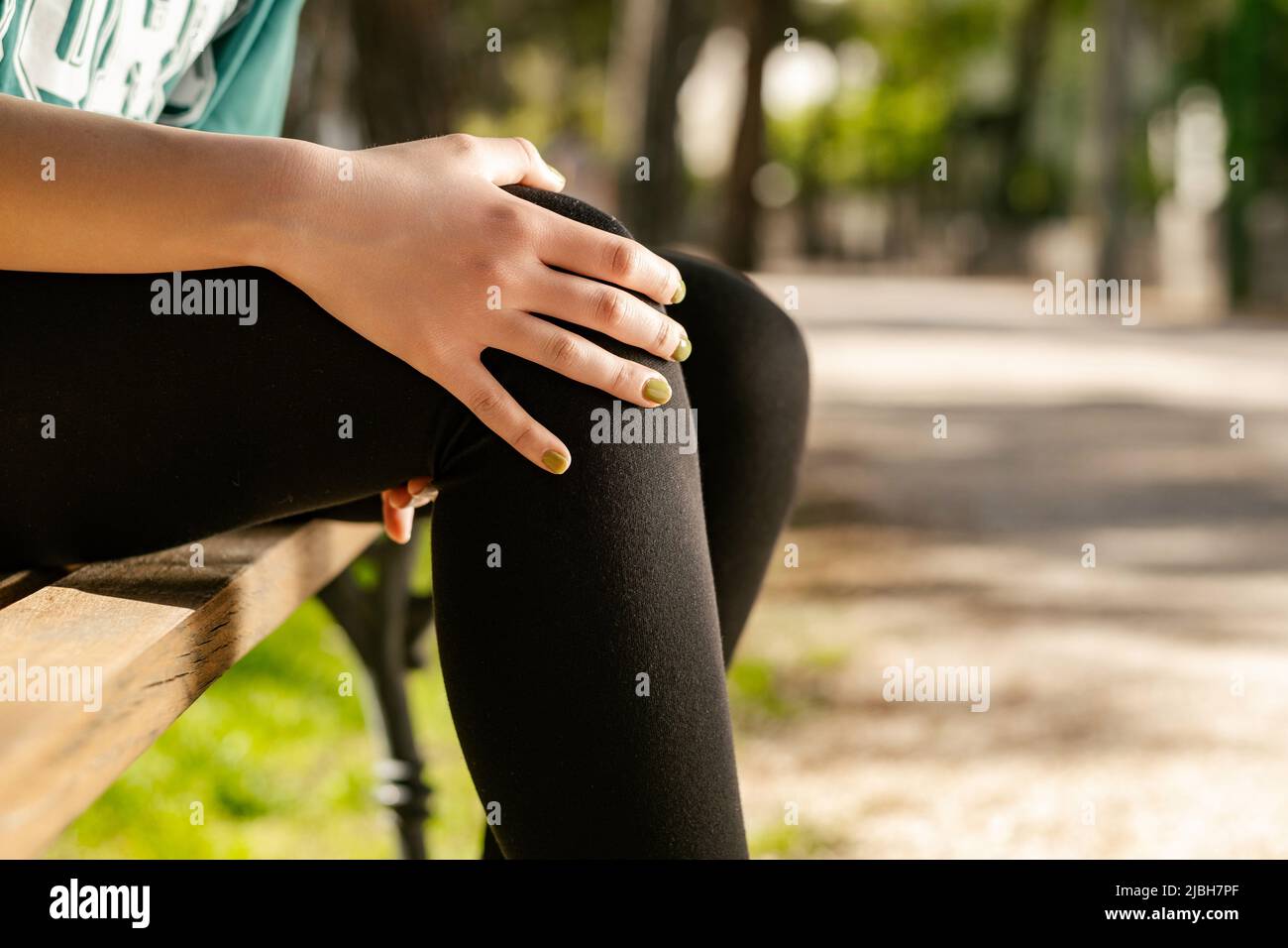 Giovane donna caucasica che indossa il tee verde in piedi sul parco della città, crampi all'aperto in gamba e sentire dolore, toccando la sua caviglia ginocchio ferita dolorosa all'esterno di poppa Foto Stock