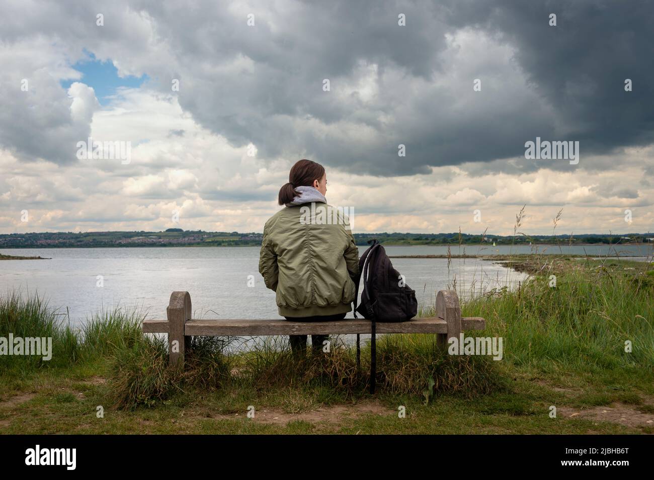 Vista posteriore di una donna seduta su una panchina da sola guardando la vista attraverso l'acqua, cielo tempestoso. Foto Stock