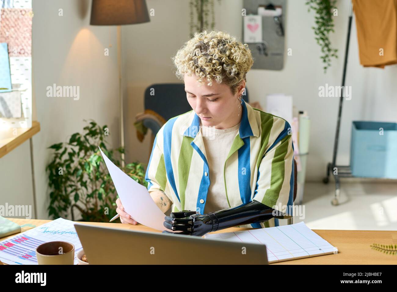 Giovane economista femminile con protesi per braccia che lavora con documenti finanziari mentre si siede sul posto di lavoro di fronte al portatile a casa Foto Stock