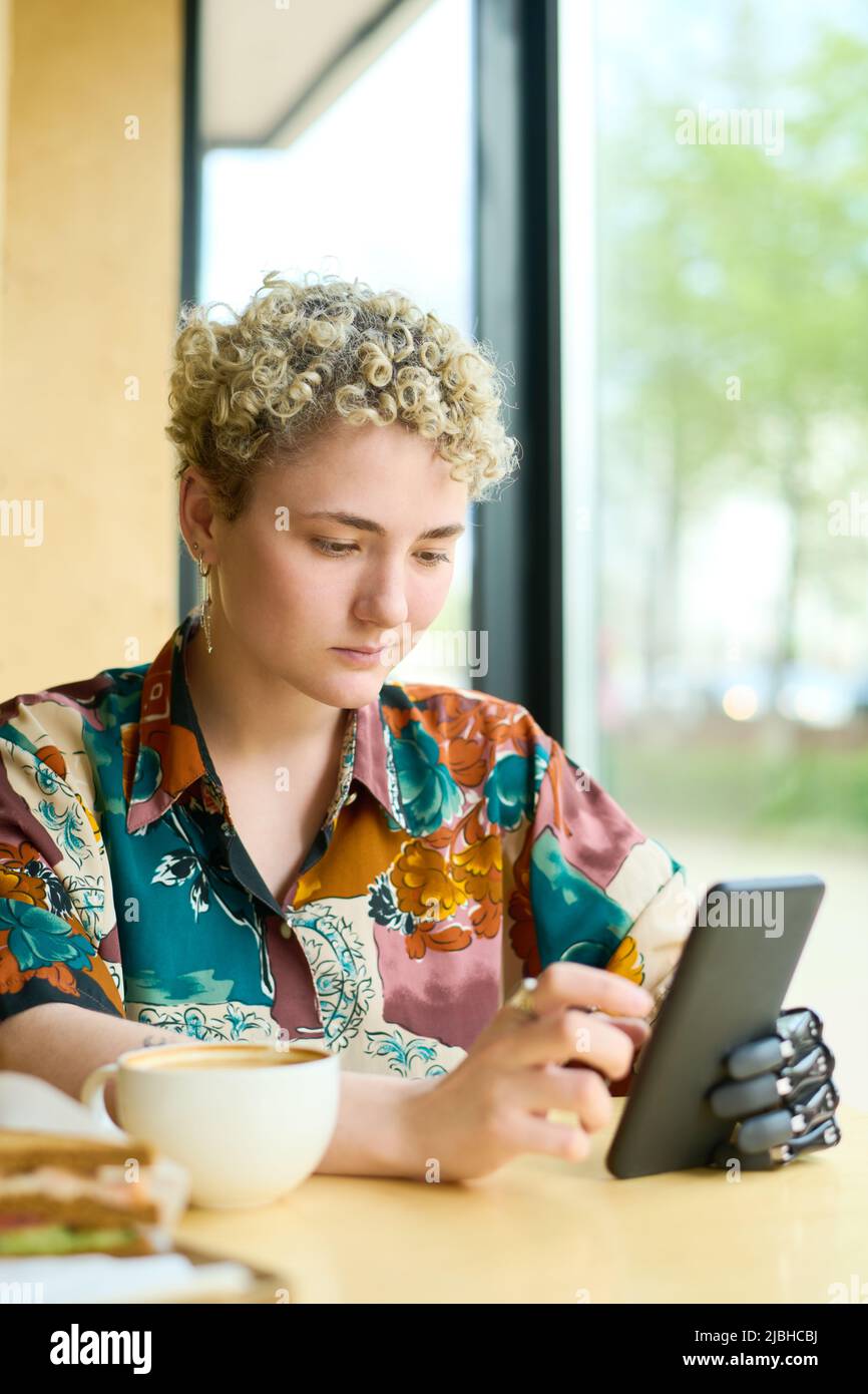 Giovane donna con braccio parziale che scorre attraverso i video online nel suo smartphone mentre si rilassa in un accogliente bar e si fa un caffè Foto Stock